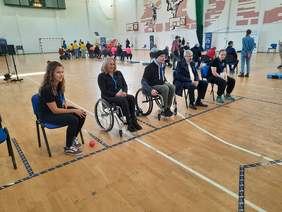 Pokaż zdjęcie: Na sali gimnastycznej w rzędzie siedzą osoby. Przy ich stopach leżą kule w czerwonych i niebieskich kolorach. Niektórzy siedzą na krzesłach, a inni na wózkach inwalidzkich. 