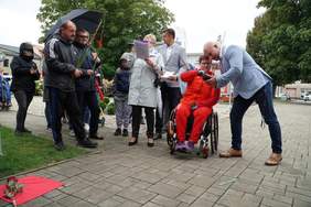 Pokaż zdjęcie: zdjęcie w plenerze w głównym miejscu widać Lucynę Kornobys siedzi na wózku obok pochyla się nad nią Bartłomiej Bonk. Po lewej stronie stoją osoby trzymają w ręku duży kwiat w kolorze czerwonym, jedna z osób trzyma mikrofon z notatkami i prze