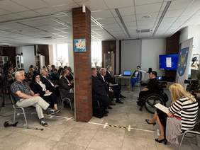 Pokaż zdjęcie: Przestronne pomieszczenie w którym około 30 osób siedzi na krzesłach ustawionych w rzędach. Na przeciwko nich jest ustawiony mężczyzna na wózku inwalidzkim.