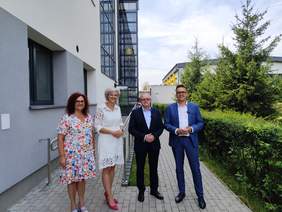 Pokaż zdjęcie: Cztery osoby pozują do zdjęcia przy podjeździe dla wózków inwalidzkich prowadzącym do windy zewnętrznej wybudowanej przy budynku szkoły