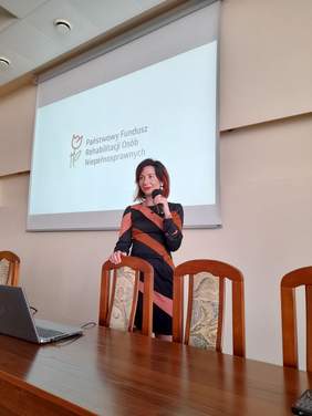 Pokaż zdjęcie: Za stołem, z mikrofonem w ręku stoi kobieta w brązowo czarnej sukience.