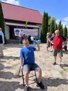 Pokaż zdjęcie: Mężczyzna w niebieskiej koszulce podnosi niebieskie hantle, siedząc na krześle przed budynkiem. W tle widać ludzi i baner zawodów sportowych dla osób niepełnosprawnych.