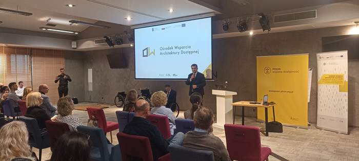 Pokaż zdjęcie: Dyrektor Oddziału Lubuskiego PFRON na tle prezentacji, obok Wiceprezes Fundacji Aktywnnej Rehabilitacji na wózku inwalidzkim, widać zgromadzonych gości siedzących na krzesłach, sala jest oświetlona, w kolorystyce ciemnej