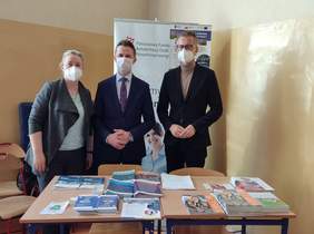 Pokaż zdjęcie: na zdjęciu w pomieszczeniu stoją trzy osoby przy stoisku z ulotkami i przygotowanymi informacjami o ofertach PFRON, z tyłu znajduje się baner z logo Funduszu