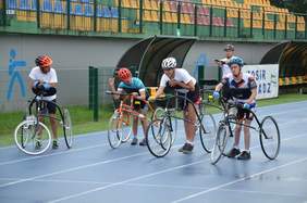 Pokaż zdjęcie: na zdjęciu otwarta przestrzeń stadionu w Sieradzu, 4 zawodników na 3 kołowych rowerach przygotowuje się do staru. 