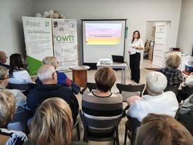 Pokaż zdjęcie: Kobieta stoi przed ekranem rzutnika, na którym wyświetlana jest prezentacja. Przed nią siedzą uczestnicy spotkania.