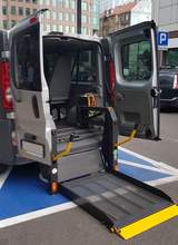 Tył samochodu typu bus, przystosowanego do przewozu osób z niepałnosprawnością. Tylna klapa jest otwarta i tworzy jednocześnie platformę do wjeżdżania dla wózków