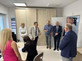 Pokaż zdjęcie: W pokoju biurowym po lewej stronie są trzy kobiety, jedna siedzi przy biurku. Po prawej stronie stoi pięciu mężczyzn i rozmawia ze sobą. Za nimi wielka szafa biurowa z jasnego drewna.