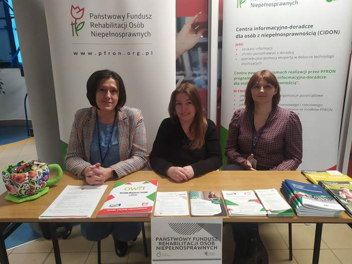 Pokaż zdjęcie: Za stołem na którym leżą ulotki, siedzą trzy kobiety. Za nimi stoją rollupy Państwowego Funduszu Rehabilitacji Osób Niepełnosprawnych oraz CIDON