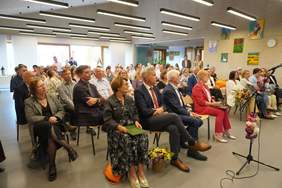 Pokaż zdjęcie: Duża grupa kobiet i mężczyzn siedzi w sali konferencyjnej na krzesłach ustawionych w kilku rzędach. W pomieszczeniu możemy dostrzec różne obrazy zawieszone na ścianie, które zostały wykonane przez osoby z niepełnosprawnością. Z przodu sali ustawiony został udekorowany kwiatami mikrofon. Z tyłu sali widoczne są duże okna, a pod sufitem podwieszane podłużne lampy.