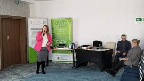 Pokaż zdjęcie: Anna Okupińska przedstawia na prezentacji funkcjonowanie OWIT. Ubrana w różową marynarkę. W prawej strony widac rollup PFRON i OWiT oraz dostępny na prezentacji sprzęt wspomagający. 