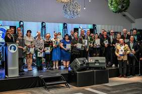 Pokaż zdjęcie: Grupa dwudziestu ośmiu osób stoi na scenie. Większość z osób trzyma w dłoniach czerwone róże oraz teczki z logotypem Zakładu Aktywności Zawodowej w Słupcy. Jedna z kobiet stoi przy mównicy. W tle są cztery reflektory, a nad sceną dekoracyjne kule.