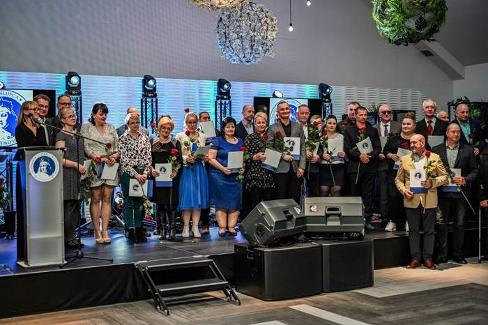 Pokaż zdjęcie: Grupa dwudziestu ośmiu osób stoi na scenie. Większość z osób trzyma w dłoniach czerwone róże oraz teczki z logotypem Zakładu Aktywności Zawodowej w Słupcy. Jedna z kobiet stoi przy mównicy. W tle są cztery reflektory, a nad sceną dekoracyjne kule.