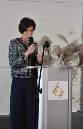 Pokaż zdjęcie: Kobieta, która trzyma w ręce mikrofon, stoi przed mównicą z logotypem Środowiskowego Domu Samopomocy. W tle dekoracja wykonana z wysokich traw ozdobnych.