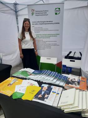 Pokaż zdjęcie: Na zdjęciu znajduje się stoisko wystawiennicze Łódzkiego Oddziału PFRON. Młoda kobieta stoi za stołem, na którym leżą różne informatory, poradniki, broszury informacyjne i ulotki. 