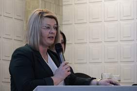 Pokaż zdjęcie: Kobieta blondynka w okularach trzyma w ręku mikrofon i przemawia.