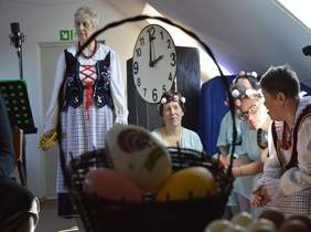 Pokaż zdjęcie: po środku koszyk wiklinowy z pisankami obok stoją osoby głównie kobietu przebrane w stroje ludowe w tle duży zegar jako scenografia do sztuki wystawianej przez uczestników WTZ