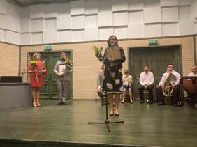 Pokaż zdjęcie: Przy mikrofonie, z dyplomem i słonecznikiem w dłoni stoi kobieta  w okularach i w sukience w białe kwiaty. W tle dwóch innych laureatów i orkiestra składająca się z osób niepełnosprawnych