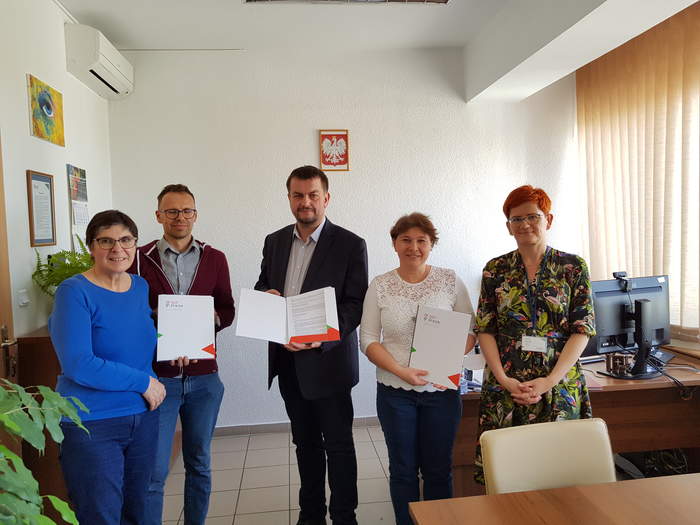 Pokaż zdjęcie: Obok siebie stoją w gabinecie Dyrektora Oddziału PFRON w Zielonej Górze osoby podpisujące umowę, wszyscy są uśmiechnięci, Dyrektor trzyma w ręku egzemplarz umowy