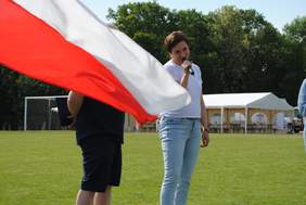 Pokaż zdjęcie: Na boisku stoją dwie osoby mężczyzna i kobieta. Kobieta [przemawia przez mikrofon, mężczyzna przesłonięty jest powiewającą flagą