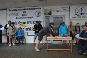 Pokaż zdjęcie: Mężczyzna oparty o stół czeka na uderzenie wiosłem. W tle dużo uśmiechniętych ludzi, którzy przyglądają się pasowaniu i mężczyzna, który zamachnął się wiosłem do uderzenia. 