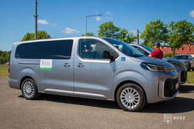 Pokaż zdjęcie: Srebrny samochód przystosowany do przewozu osób z niepełnosprawnościami stoi na parkingu w słoneczny dzień. Na pojeździe widoczne są oznaczenia PFRON oraz symbol dostępności dla osób poruszających się na wózkach.