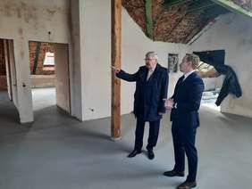 Pokaż zdjęcie: We wnętrzu budowanego domu znajduje się dwóch mężczyzn. Jeden z nich wskazuje na coś palcem. Oceniają postęp prac budowy. Panowie są ubrani w garnitury. 