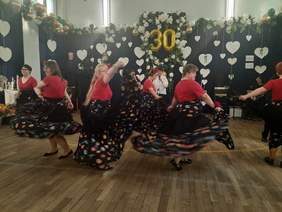 Pokaż zdjęcie: Na środku sali kobiety tańczą w stylu cygańskim. Mają czerwone bluzki i piękne długie kolorowe spódnice. Wszystkie robią piruety. 