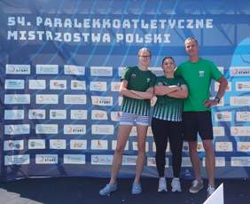 Pokaż zdjęcie: Trzy osoby pozują do zdjęcia na tle ścianki pięćdziesiątych czwartych Paralekkoatletycznych Mistrzostw Polski