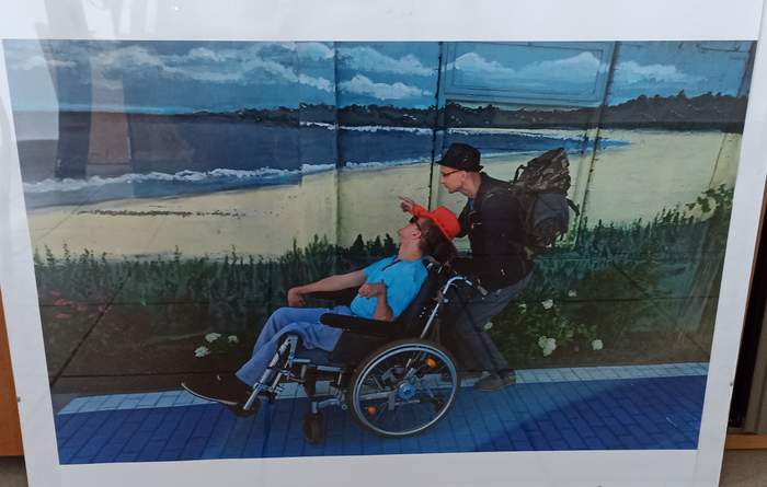 Pokaż zdjęcie: Osoba na wózku i opiekun tej osoby na molo plaży, ubrani w stroje letnie