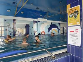 Pokaż zdjęcie: Dwie kobiety i czworo mężczyzn pływa w basenie. Ściany pomieszczenia wyłożone są różnokolorowymi płytkami, a jednej z nich powieszone są plakaty promujące Dni Sportu Adaptowanego i Zdrowia oraz informujące o wysokości dofinansowania ze środków PFRON.