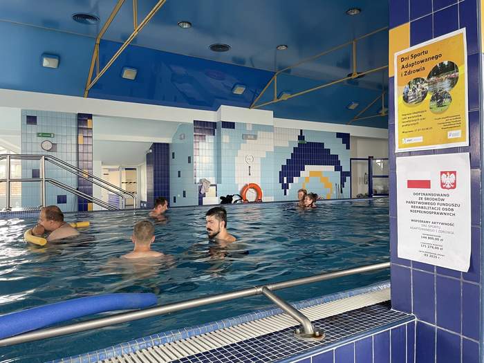 Pokaż zdjęcie: Dwie kobiety i czworo mężczyzn pływa w basenie. Ściany pomieszczenia wyłożone są różnokolorowymi płytkami, a jednej z nich powieszone są plakaty promujące Dni Sportu Adaptowanego i Zdrowia oraz informujące o wysokości dofinansowania ze środków PFRON.