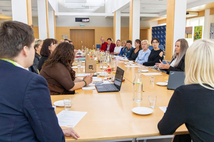 Pokaż zdjęcie: Grupa kilkunastu osób siedzi przy połączonych ze sobą stołach, na których ustawione zostały napoje oraz różne owoce w formie poczęstunku. Każdy z uczestników ma przed sobą program spotkania.