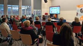 Pokaż zdjęcie: po lewej stronie siedzą osoby w dużej ilości i słuchają prelegenta seminarium po środku biurko na górze telebim  z prezentacją. Za biurkiem siedzi osoba i prezentuje swoje wystąpienie