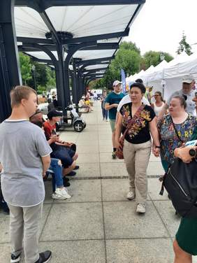 Pokaż zdjęcie: Na Starym Rynku w Łodzi widać wielu spacerowiczów. Są wśród nich osoby z niepełnosprawnościami. Po prawej stronie znajdują się różne stoiska dedykowane osobom z niepełnosprawnościami.   