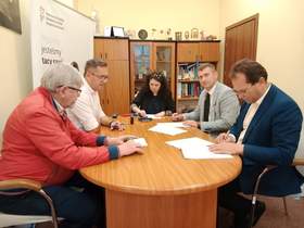 Przy owalnym stole siedzi kobieta i czterech mężczyzn. Podpisują umowę. Na stole leżą różne dokumenty, telefony, pieczątki i długopisy. 