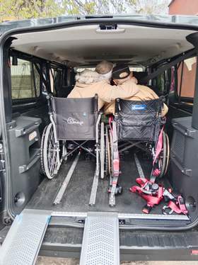 We wnętrzu samochodu dostosowanego do przewozu osób niepełnosprawnych znajduje się dwoje starszych ludzi. Siedzą na wózkach inwalidzkich i przytulają się do siebie. Widać ich siedzących tyłem. 