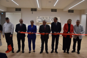 Pokaż zdjęcie: Grupa siedmiu osób stoi w sali i przecina czerwoną wstęgę podczas uroczystego otwarcia; w tle napis „Życie to droga…” na ścianie, jasne wnętrze z drewnianą podłogą i oświetleniem sufitowym.