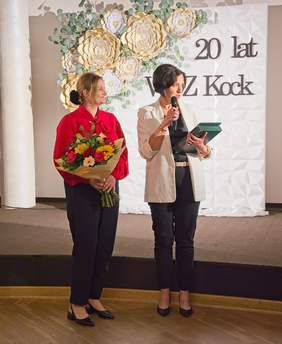 Pokaż zdjęcie: Dwie kobiety elegancko ubrane, jedna trzyma kwiaty, drugi trzyma w ręku mikrofon oraz list i grawerton.