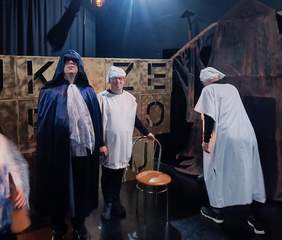 Trzy osoby w kostiumach na scenie. Jeden mężczyzna ubrany w ciemnoniebieską pelerynę z kapturem, dwóch innych w białych czepkach i fartuchach, przypominających stroje szpitalne. W tle widoczne są duże, złote sześciany z wyciętymi literami "J", "K", "R", "Z", "E".