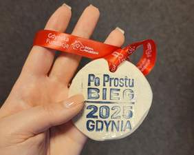 Pokaż zdjęcie: Medal z napisem „Po Prostu Bieg 2025 Gdynia” na czerwonej smyczy z logo Gdyńskiej Fundacji „Dom Marzeń”, trzymany w dłoni o jasnych paznokciach.