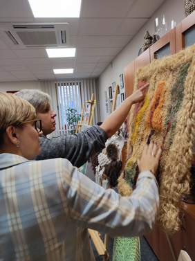 Pokaż zdjęcie: Dwie kobiety oglądają z bliska barwną tkaninę artystyczną podczas oceny konkursowych prac.