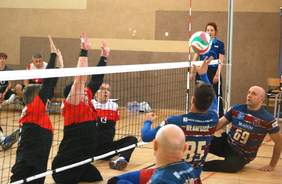 Pokaż zdjęcie: Zawodnicy innych klubów w trakcie gry na siedząco, widzimy piłkę w w powietrzu, a w tle sędzinę z gwizdkiem w ustach 