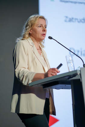 Pokaż zdjęcie: Przy mównicy stoi kobieta blondynka w białej marynarce.