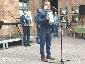 Mężczyzna w garniturze, stoi przed mikrofonem, przemawia, za nim sylwetki trzech mężczyzn którzy stoją, a z prawej strony siedzi kobieta, wszystko na tle budynku z cegły