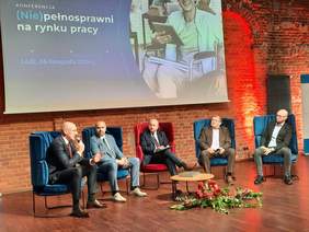 Pokaż zdjęcie: W sali siedzi pięciu mężczyzn. Jeden z panów trzyma w dłoni mikrofon, do którego przemawia. Nad mężczyznami wisi tablica multimedialna, na której wyświetlona jest nazwa konferencji: "(Nie)pełnosprawni na rynku pracy". 