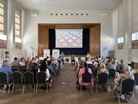 Pokaż zdjęcie: Na dużej Sali porozstawianych jest wiele krzeseł na których siedzą ludzie. Za nimi scena nad nią wisi ekran. Przed widownią stoi mężczyzna w jasnej marynarce białej koszuli, trzyma mikrofon i mówi.