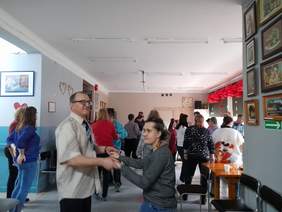 Pokaż zdjęcie: Na zdjęciu zbliżenie na parę tańczącą podczas Balu Walentynkowego, para trzyma się za ręce. W tle widać inne tańczące osoby.