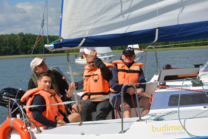 Pokaż zdjęcie: Grupa mężczyzn siedząca na pokładzie jachtu. Dwóch panów ma podniesione ręce w geście powitania.  Jeden trzyma linę. 