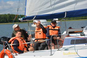 Pokaż zdjęcie: Grupa mężczyzn siedząca na pokładzie jachtu. Dwóch panów ma podniesione ręce w geście powitania.  Jeden trzyma linę. 